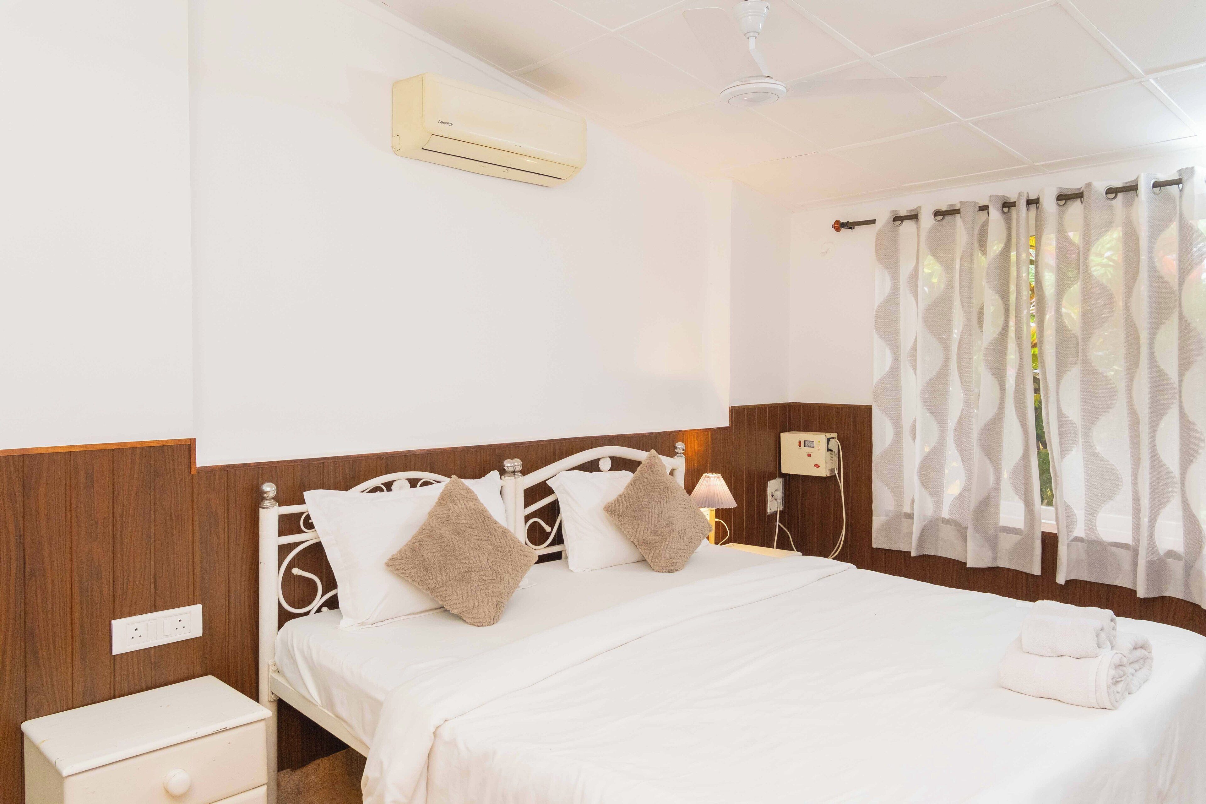 Cama bem arrumada com roupa de cama branca e duas almofadas bege, mesa de cabeceira com gaveta, ar condicionado fixado na parede e janela com cortinas estampadas.