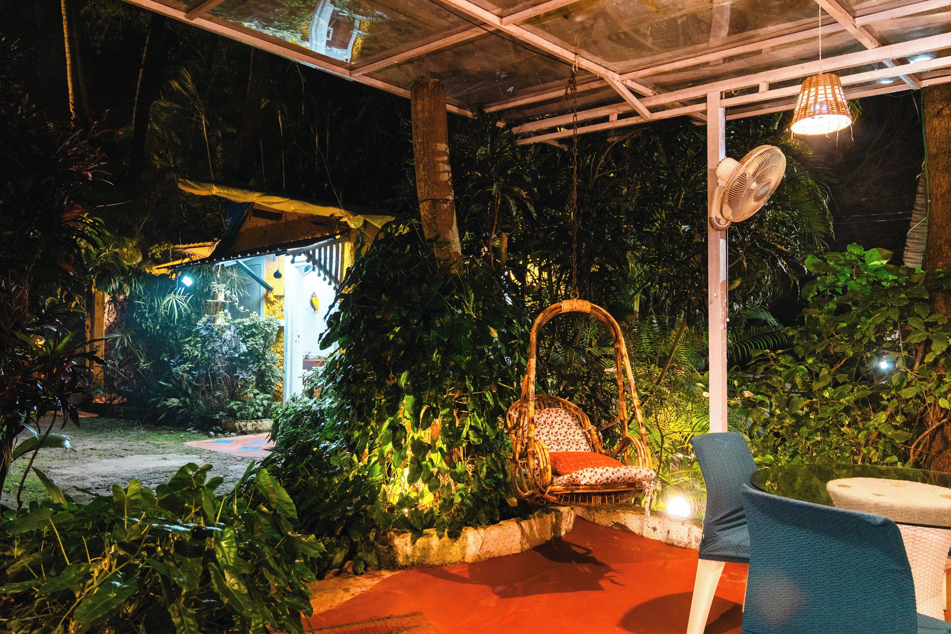 An outdoor seating area with a wicker chair and a fan, surrounded by lush greenery and a covered structure in the background.