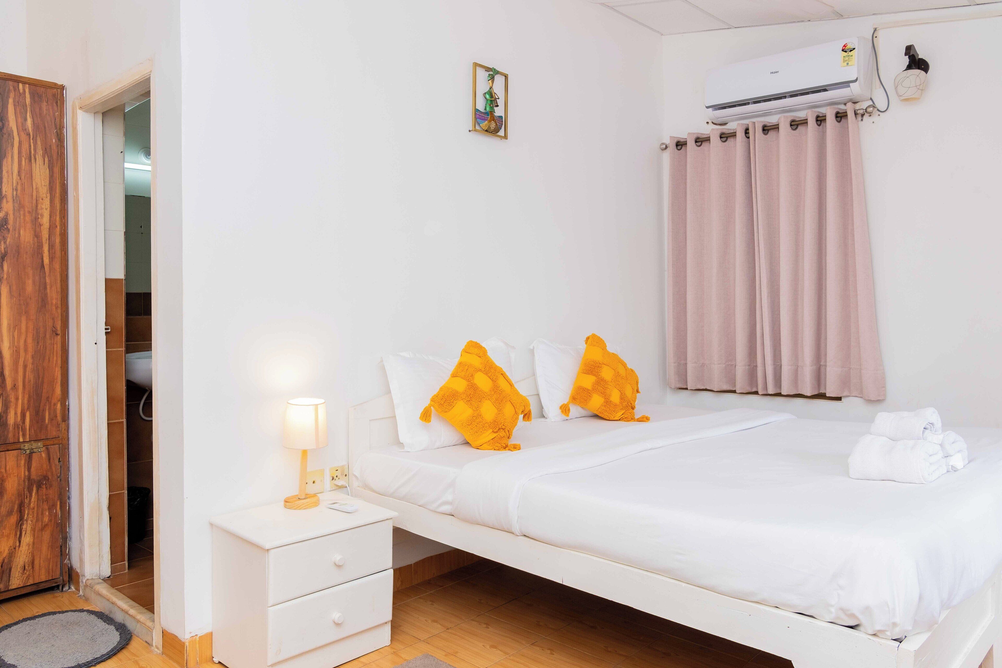 A neatly made bed with white bedding and two orange pillows, a bedside table with a lamp, a wooden wardrobe, a pink curtain, and an air conditioning unit on the wall.
