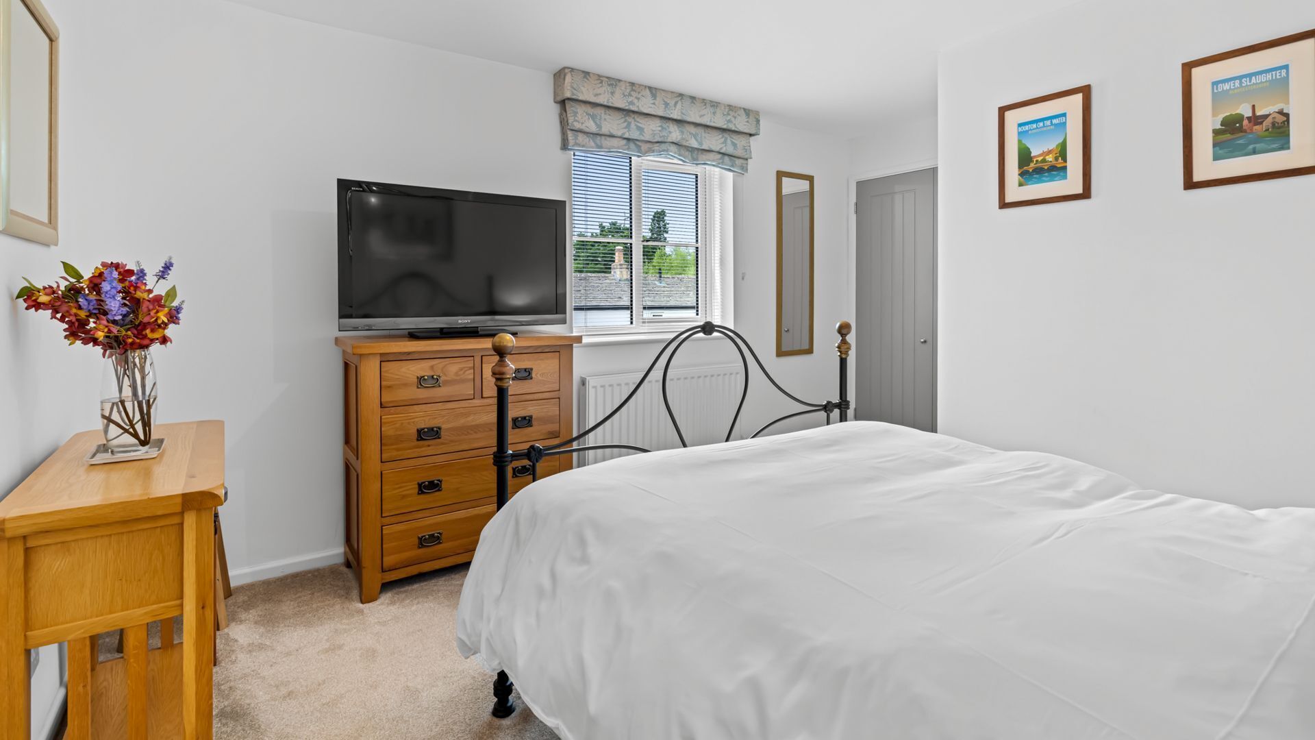 A bedroom with a bed, a wooden dresser with a television on top, a vase with flowers, and two framed pictures on the wall.