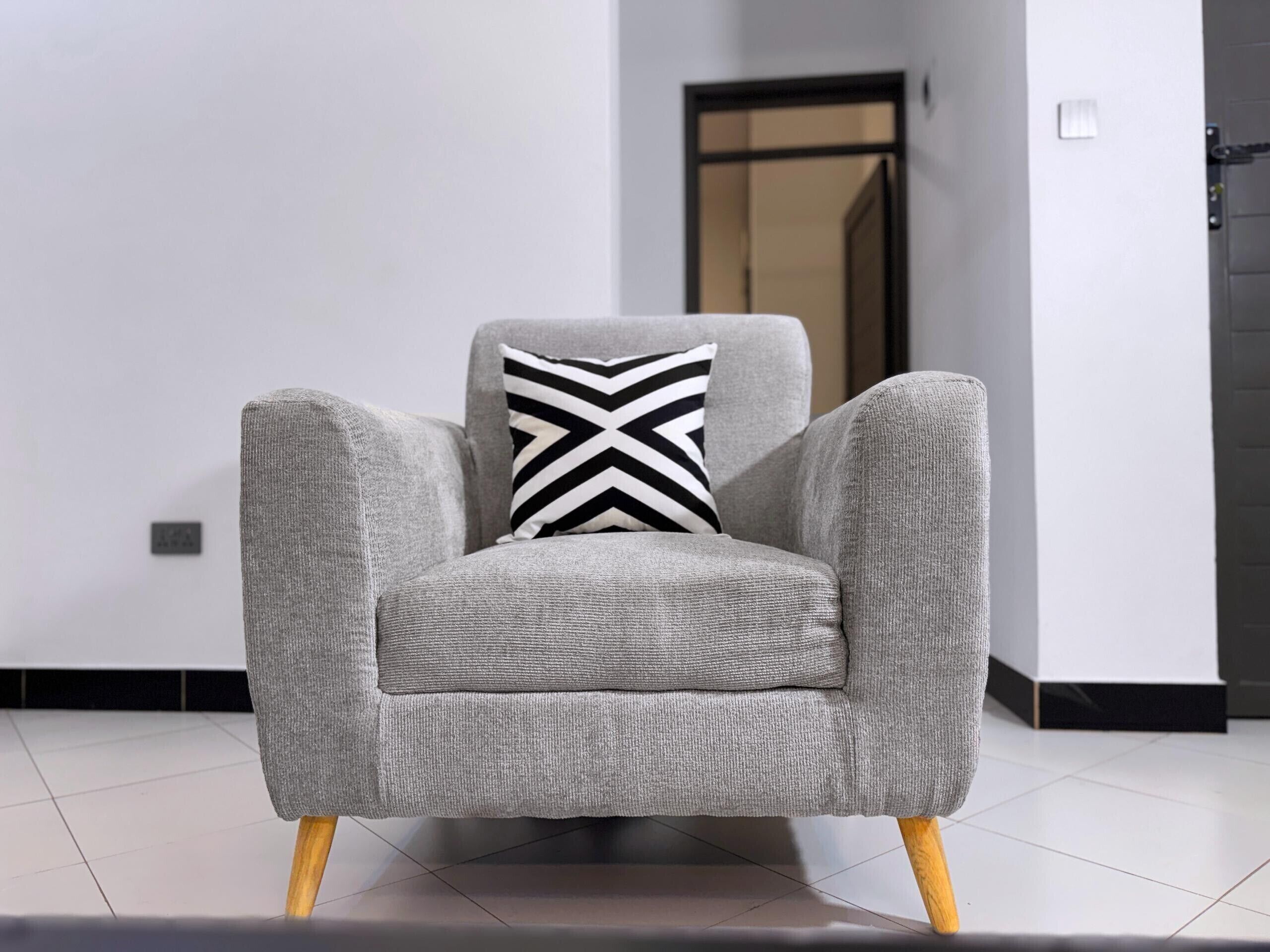 Un fauteuil gris aux pieds en bois, agrémenté d’un coussin à motifs chevrons noir et blanc, dans une pièce moderne aux murs blancs et au sol carrelé.