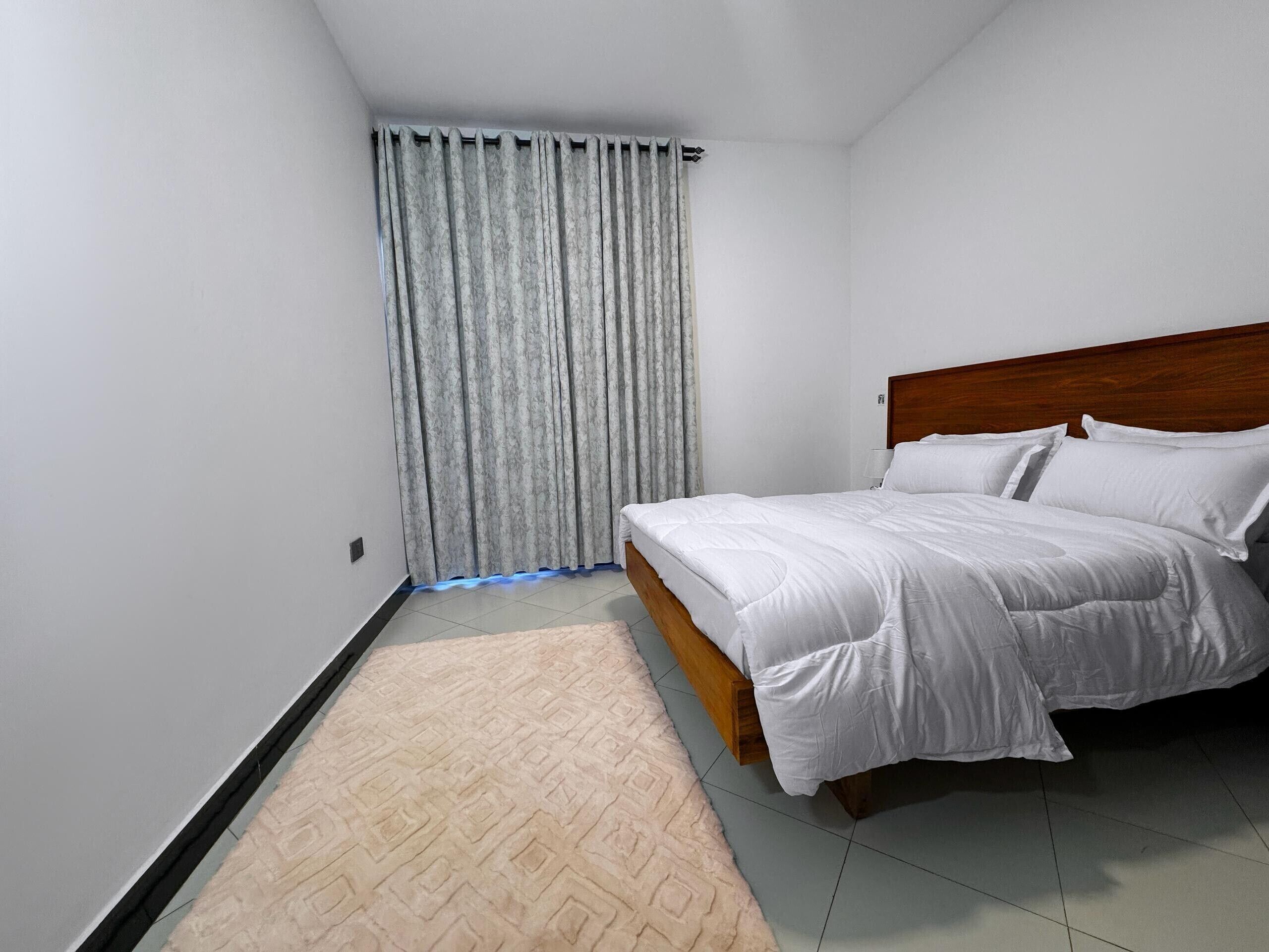 A bedroom with a wooden bed, white bedding, a patterned rug, and a window with curtains.