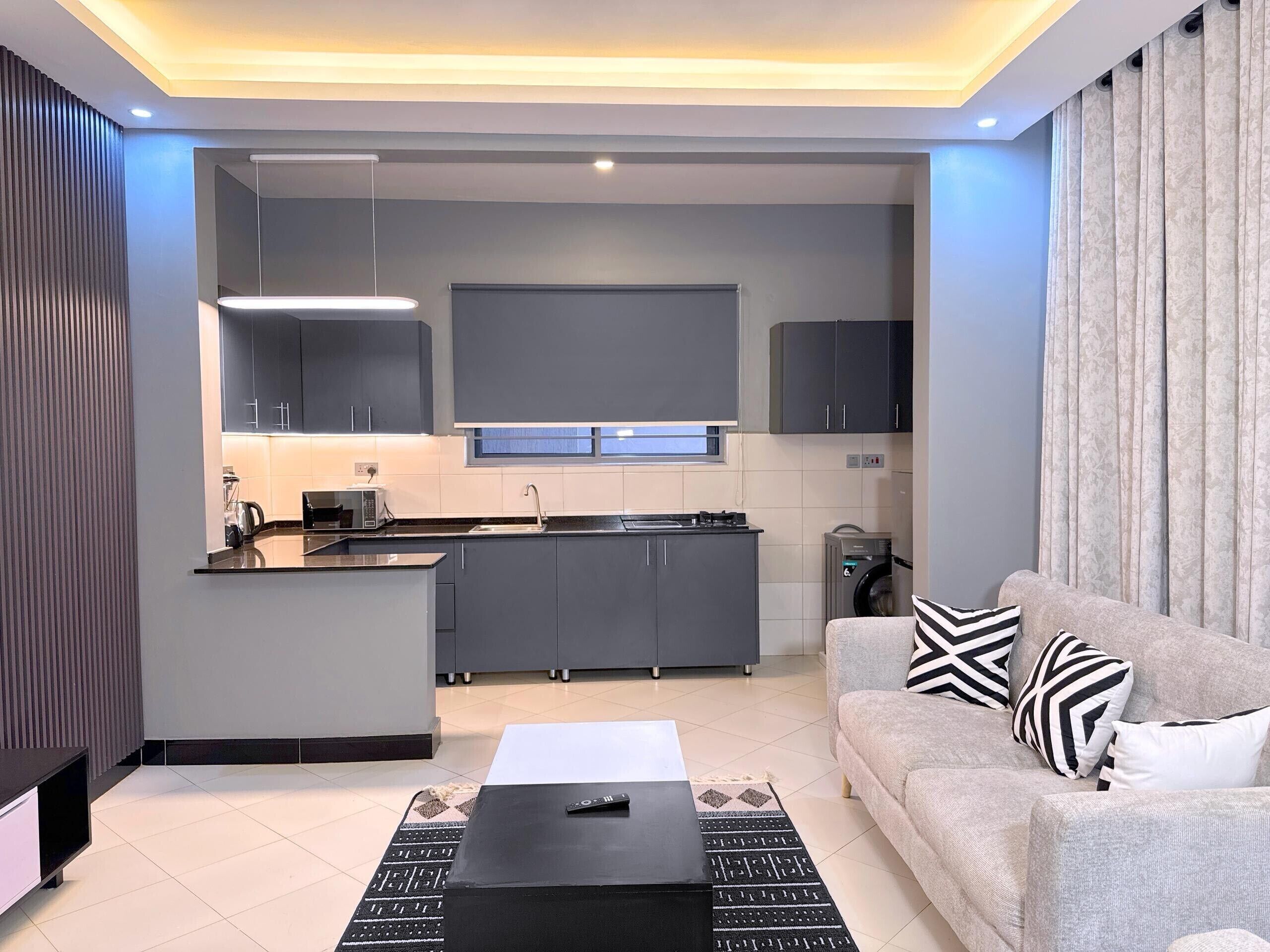 A modern living room with a grey sofa, black and white patterned rug, and a black coffee table. A kitchen with grey cabinets and a built-in island is visible in the background.
