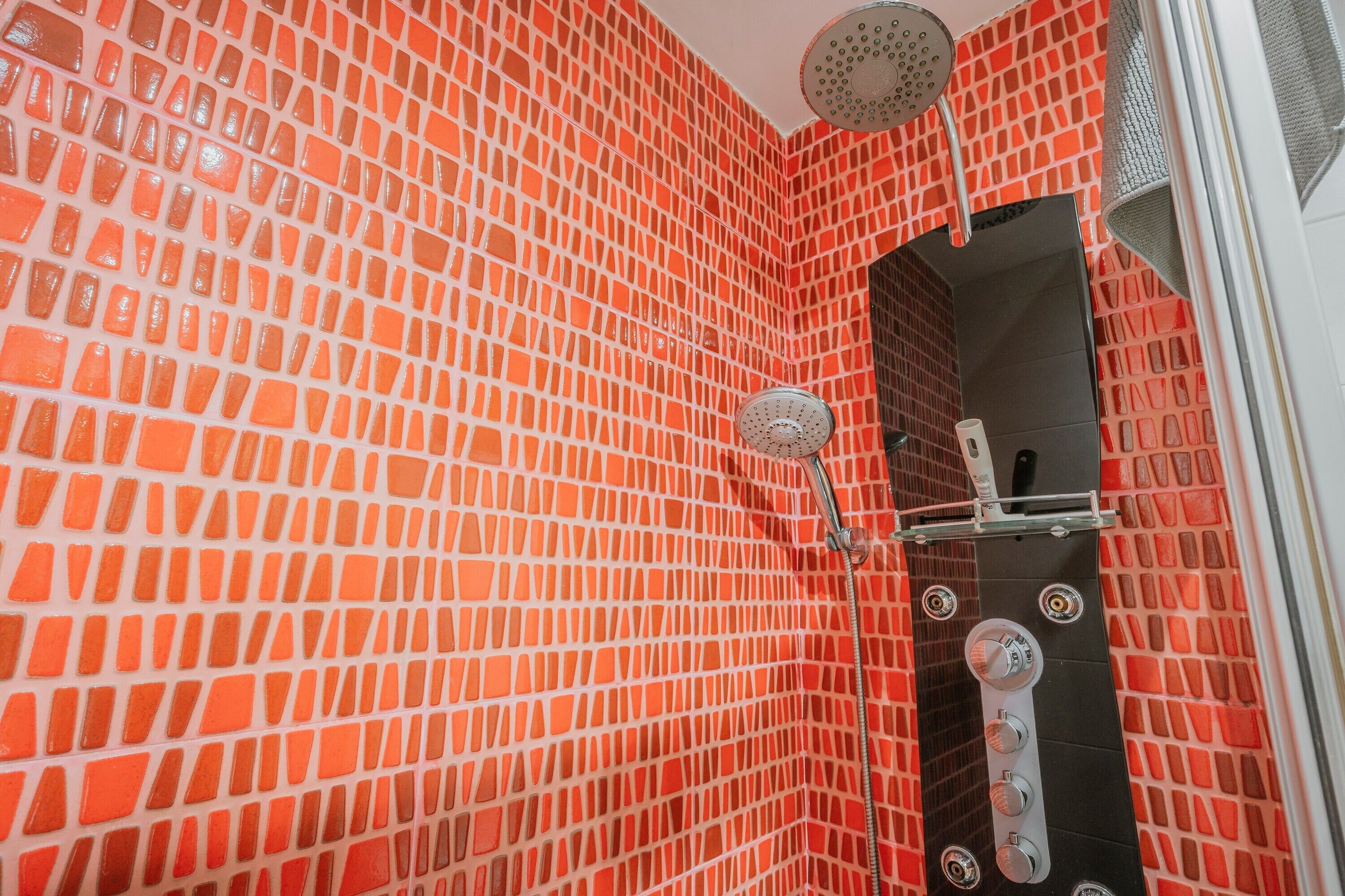 A bathroom with a red mosaic tile wall, a showerhead, and a control panel.