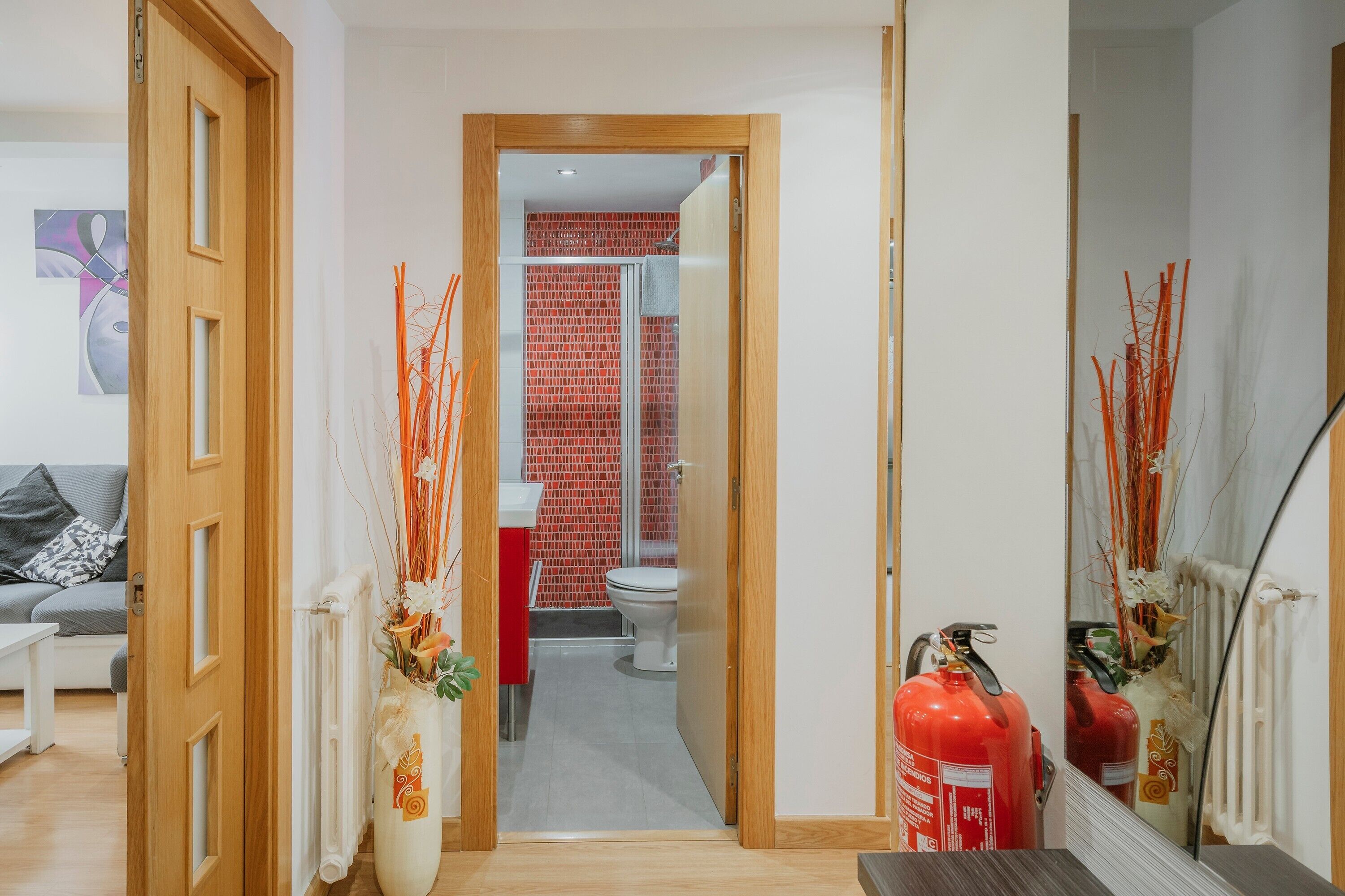 Een gang die uitkomt bij een badkamer met rode tegels, een wit toilet en een brandblusser.