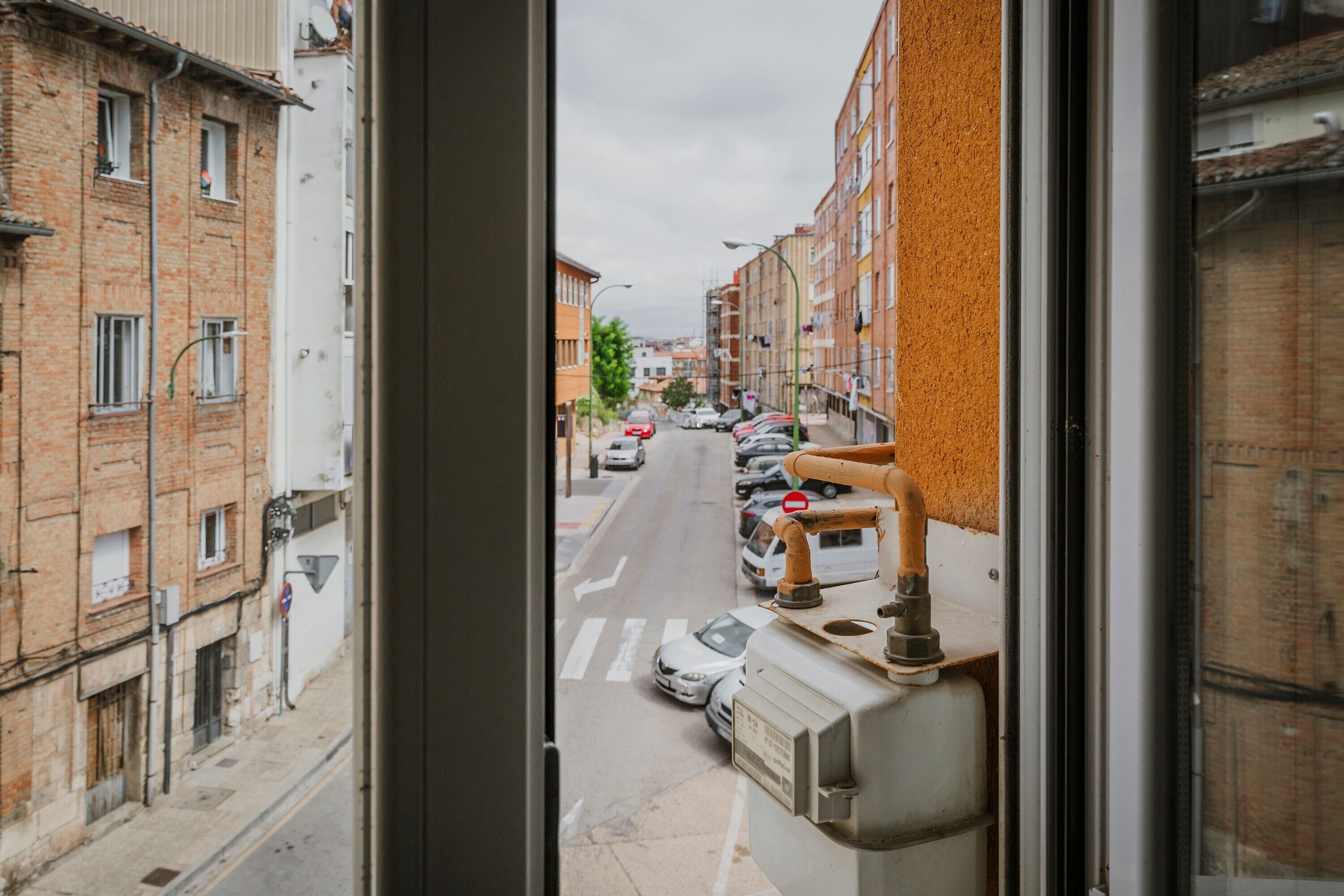 Une vue de la rue à travers une fenêtre, montrant une rangée de voitures garées, des immeubles aux façades en briques et un panneau de signalisation visible.