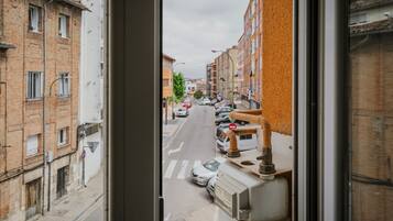 Une vue de la rue à travers une fenêtre, montrant une rangée de voitures garées, des immeubles aux façades en briques et un panneau de signalisation visible.