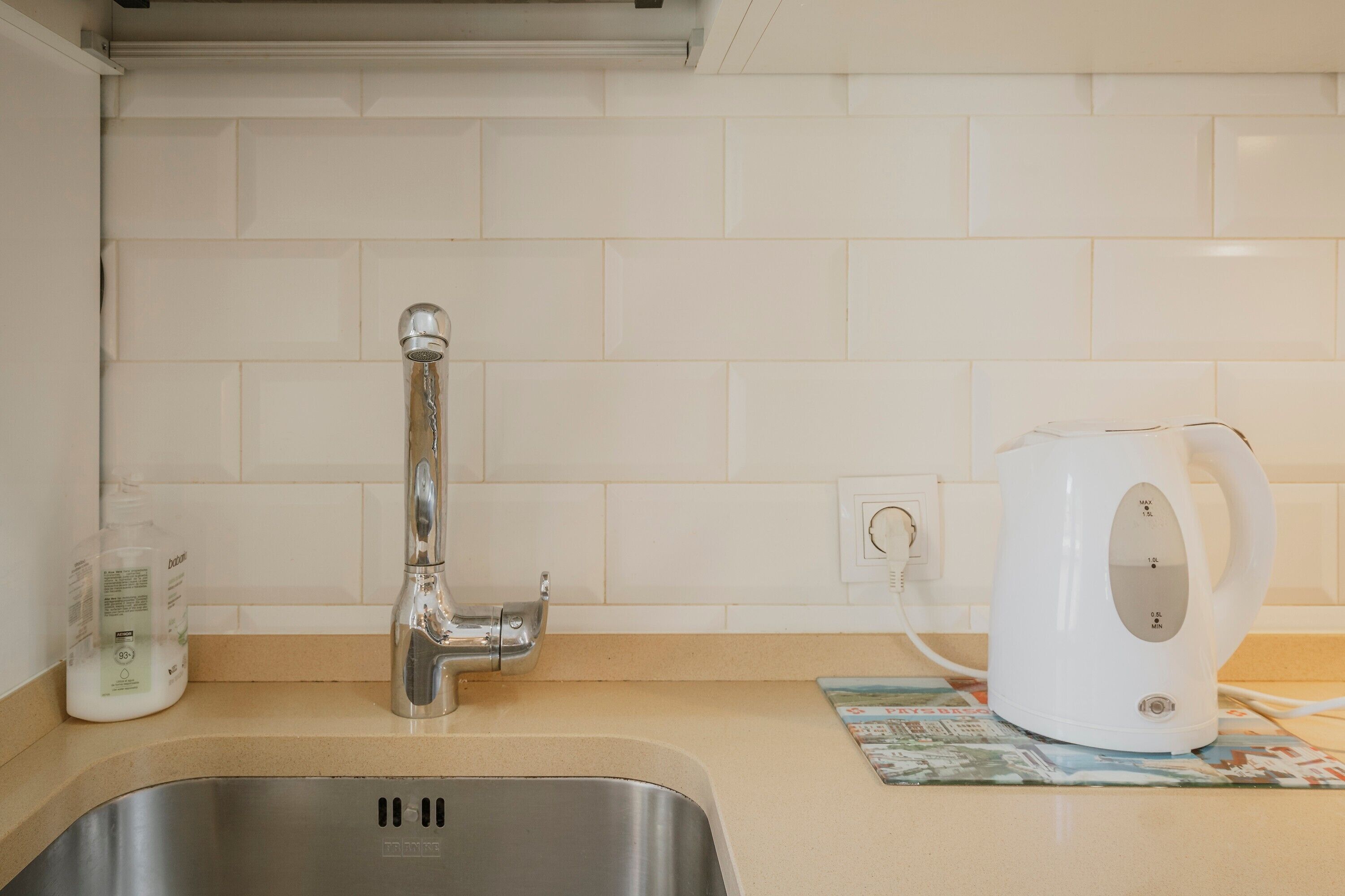 A kitchen with a stainless steel sink, a white kettle, and a bottle of dish soap on the counter.