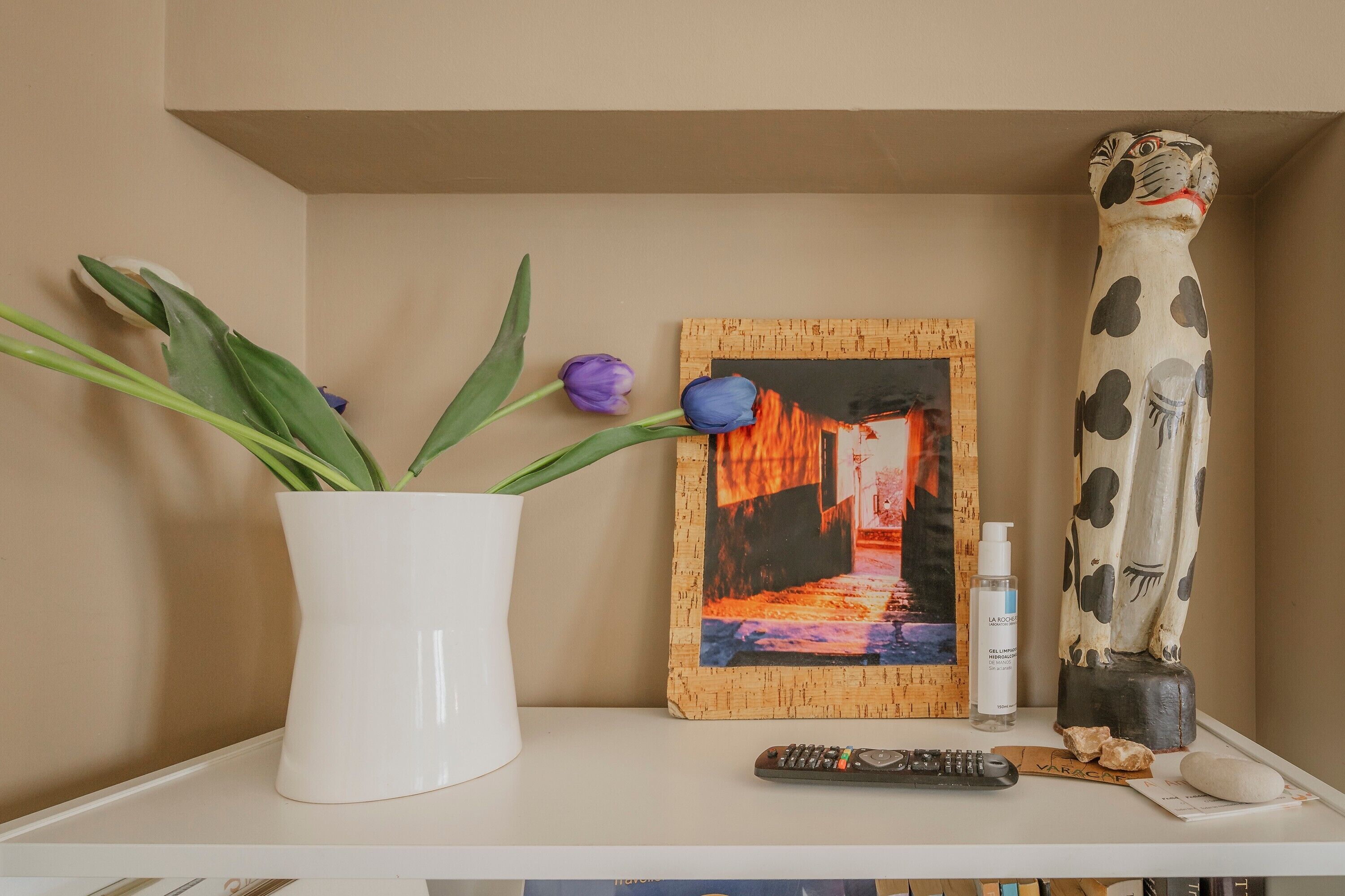 A white vase with green leaves and purple flowers, a framed picture, a remote control, a bottle, and a decorative sculpture on a shelf.