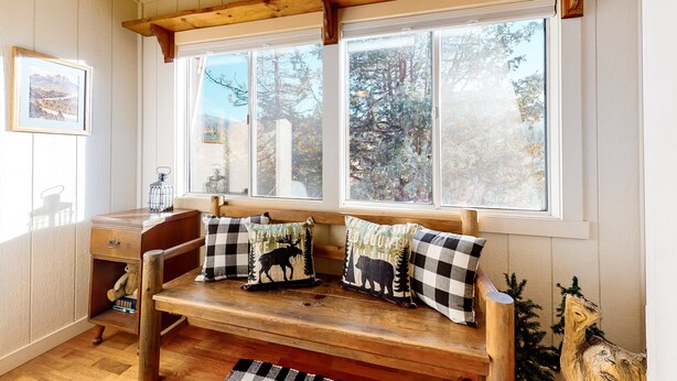 Un banco de madera con cojines a cuadros, un pequeño escritorio con un osito de peluche y una ventana con vistas a árboles.