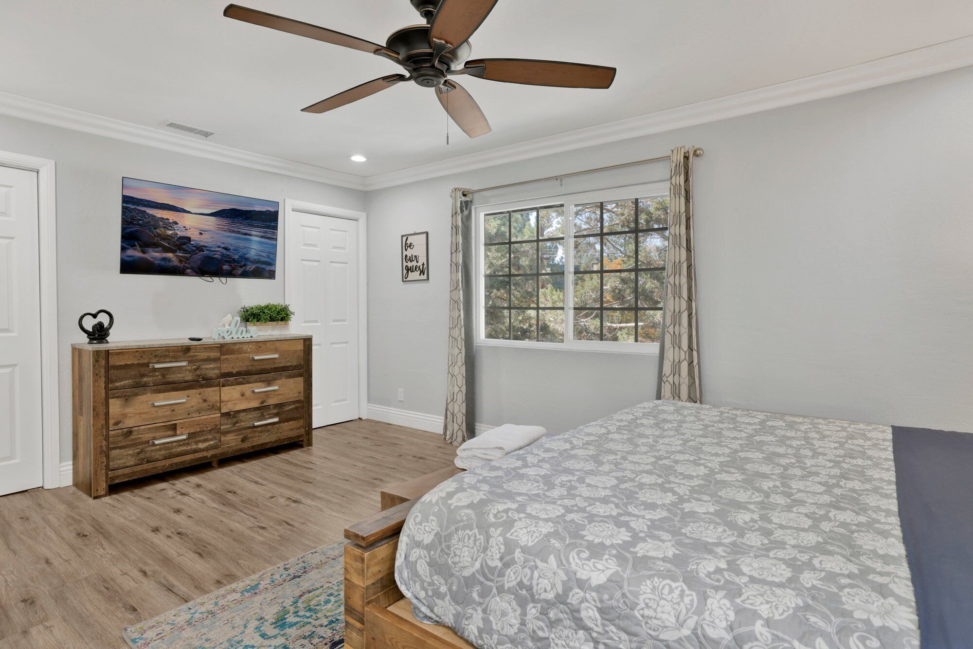 A bedroom with a bed, wooden dresser, ceiling fan, and a window with curtains.