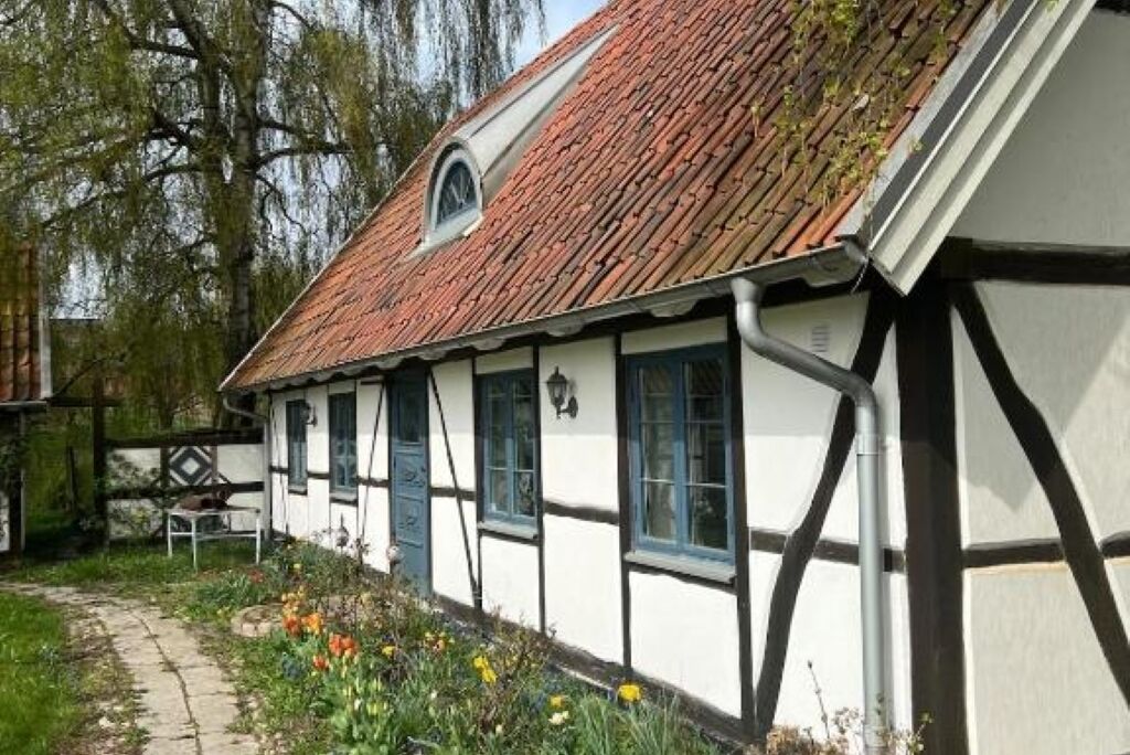 A traditional half-timbered house with a sloped roof, a small window with a triangular pediment, and a pathway leading to the entrance.