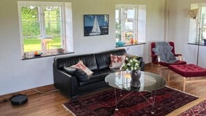 A living room with a black leather sofa, a glass coffee table, a red ottoman, and a red chair. There are two windows with white frames, a red rug, and a painting of a sailboat on the wall. - Renovated Half-timbered house in Österlen built 2006 (Skåne län)