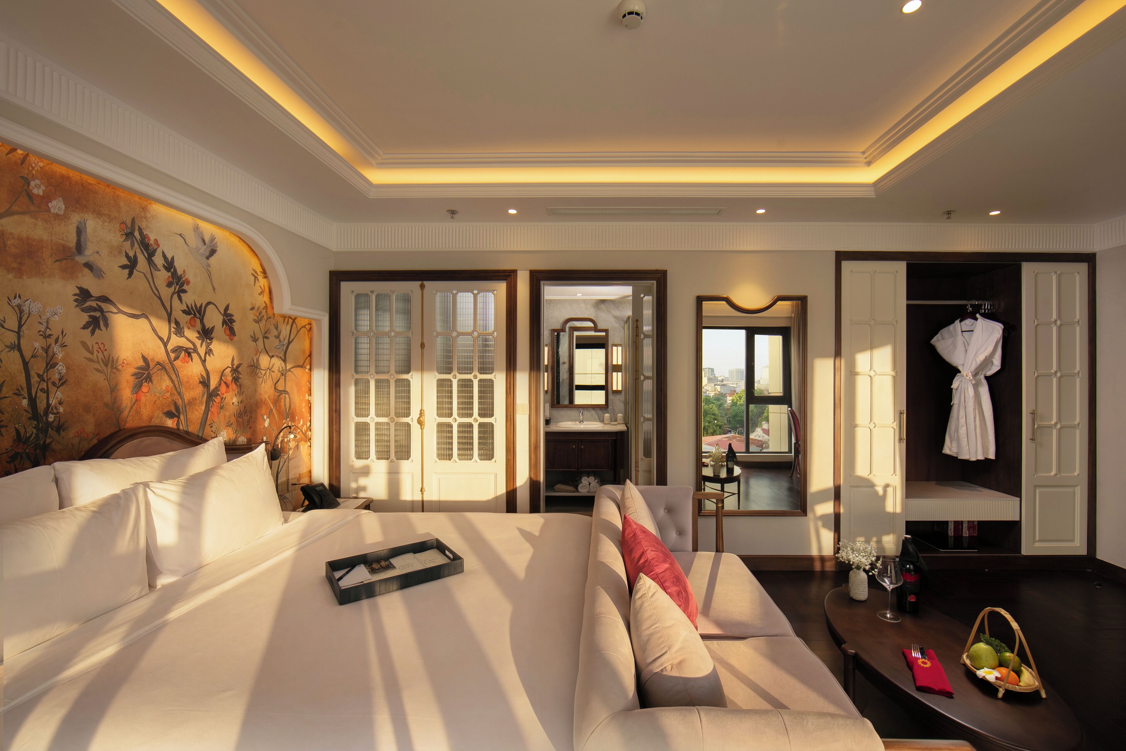 A hotel room with a large bed, a sofa, a dining table with a basket of fruit, a mirror, and a view of the city.