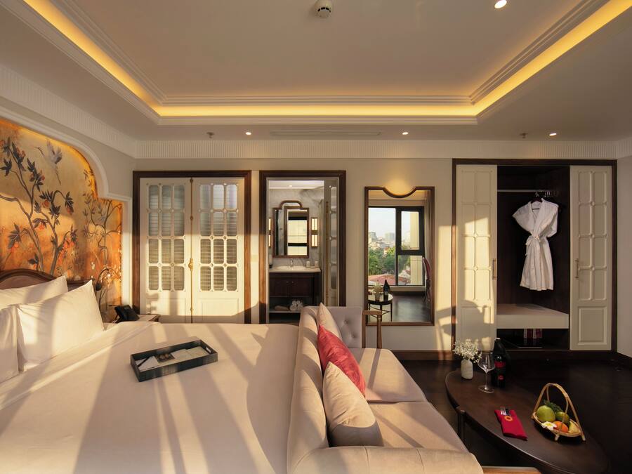 A hotel room with a large bed, a sofa, a dining table with a basket of fruit, a mirror, and a view of the city.