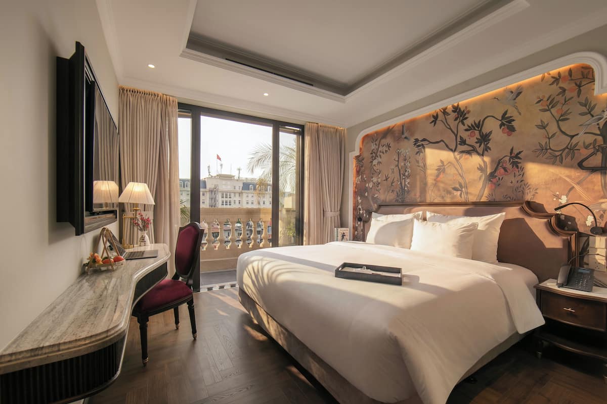 Hotelkamer met een groot bed, een bureau met een lamp, een stoel en uitzicht op de stad door een groot raam.