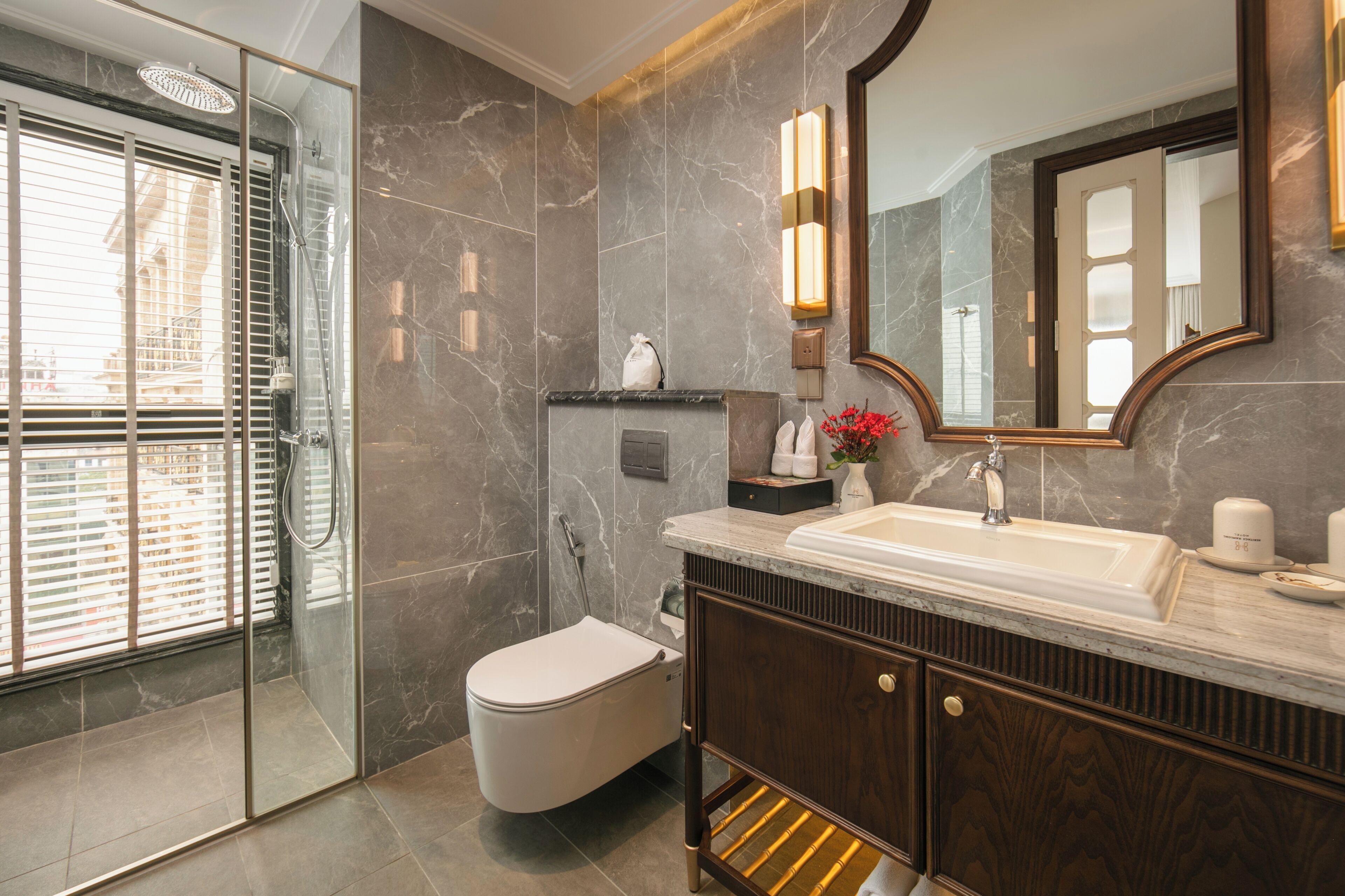 Un baño moderno con ducha envuelta en vidrio, inodoro blanco, pared con azulejos de mármol, tocador de madera y un espejo grande.