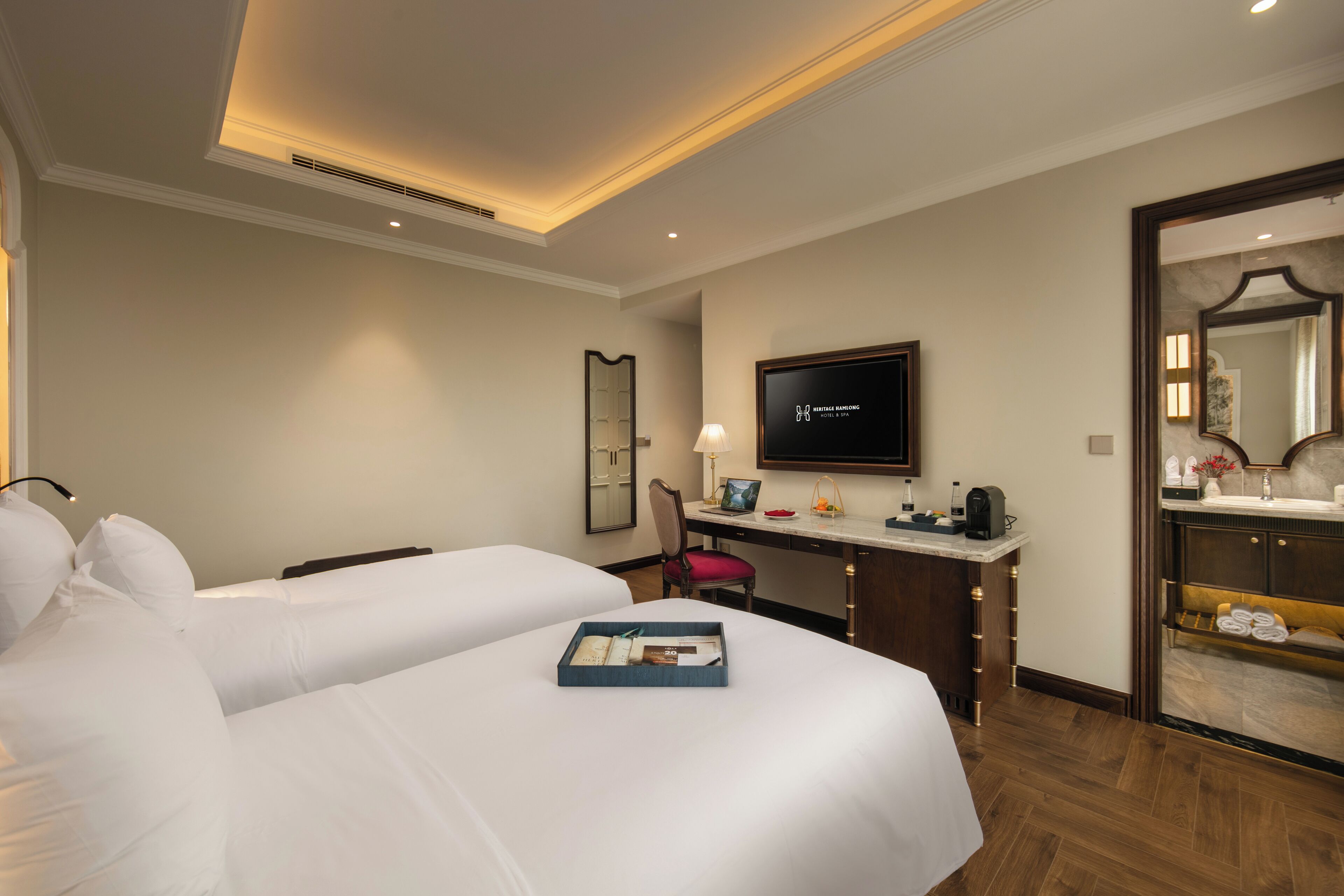 Une chambre d’hôtel avec deux lits, un bureau avec un ordinateur, une télévision et une salle de bain équipée d’un miroir et d’un porte-serviettes.