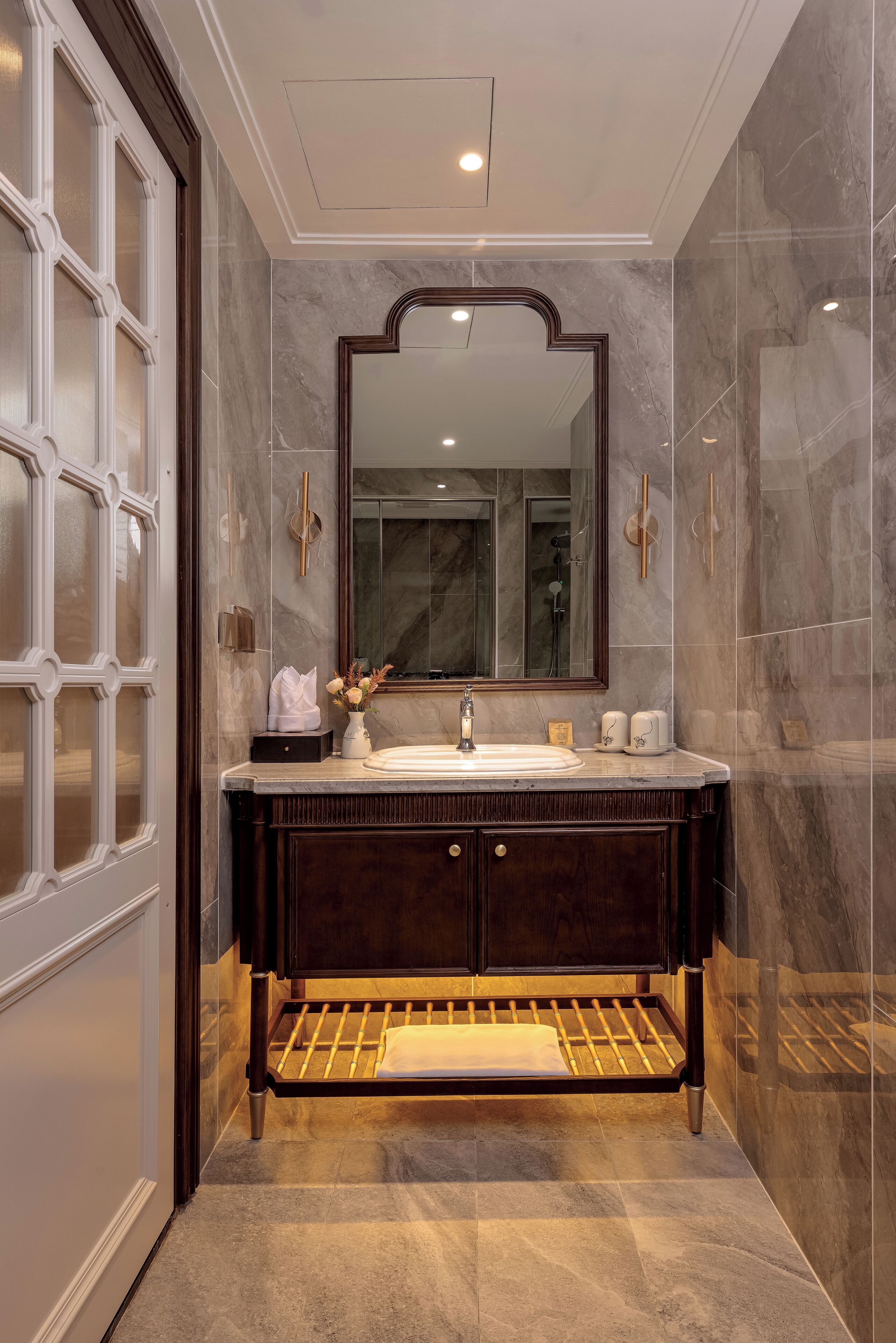 A modern bathroom with a large mirror, dark wood vanity, marble walls, and a built-in towel rack.