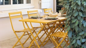 Un coin repas avec des chaises jaunes et une table dressée pour quatre personnes, une porte blanche et une fenêtre avec des stores.