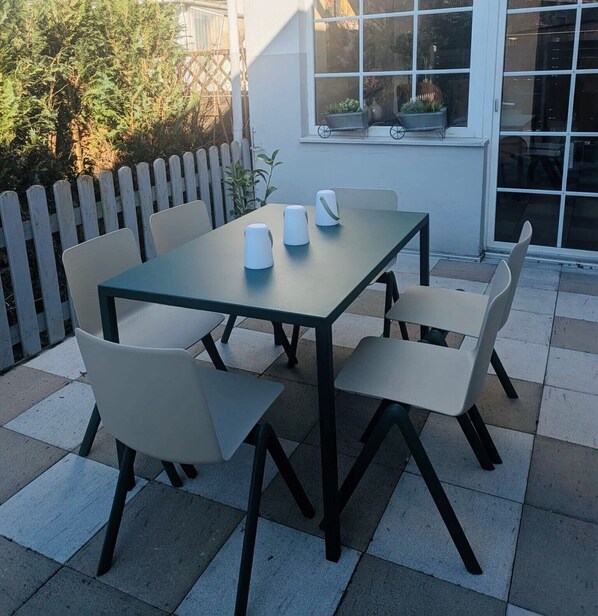 An outdoor patio with a table and chairs, a white picket fence, and a window with potted plants.