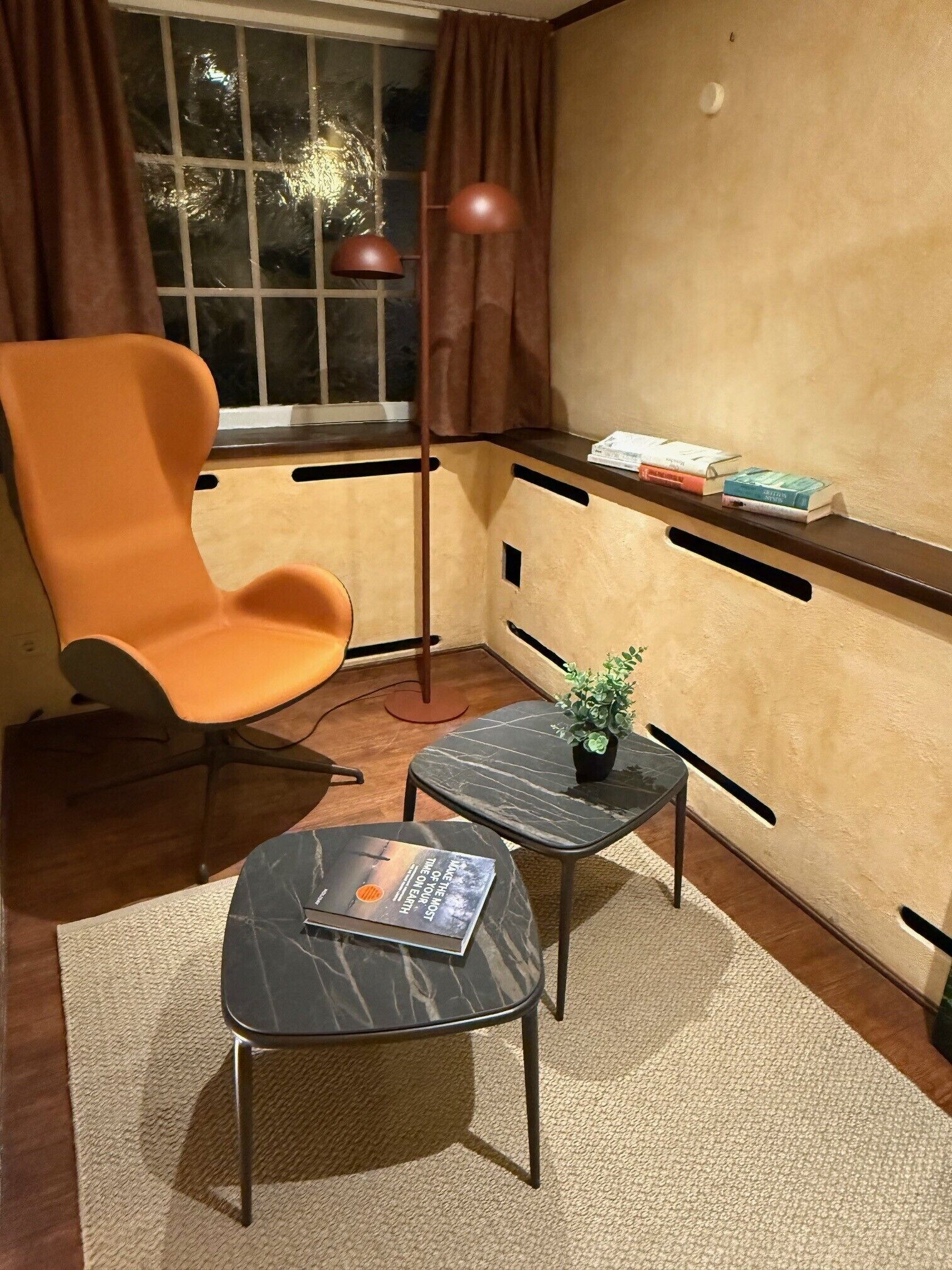 A corner room with a modern orange chair, a small black table with a book, a potted plant, and a window with curtains.