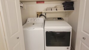 A laundry room with two stacked washing machines, a shelf with folded towels, and a sign warning against removing the washer. - Clean Spacious 3BR, ½ Mi to Hospital, Near Universities, Fast WiFi, Pet-Friendly (Princeton)
