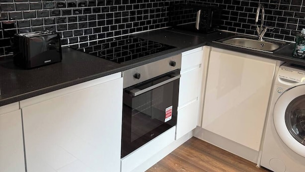 A compact kitchen with white cabinets, a black tiled backsplash, a built-in oven, a washing machine, and a sink. - Nice 1-bedroom apartment in Milton Keynes (Milton Keynes)