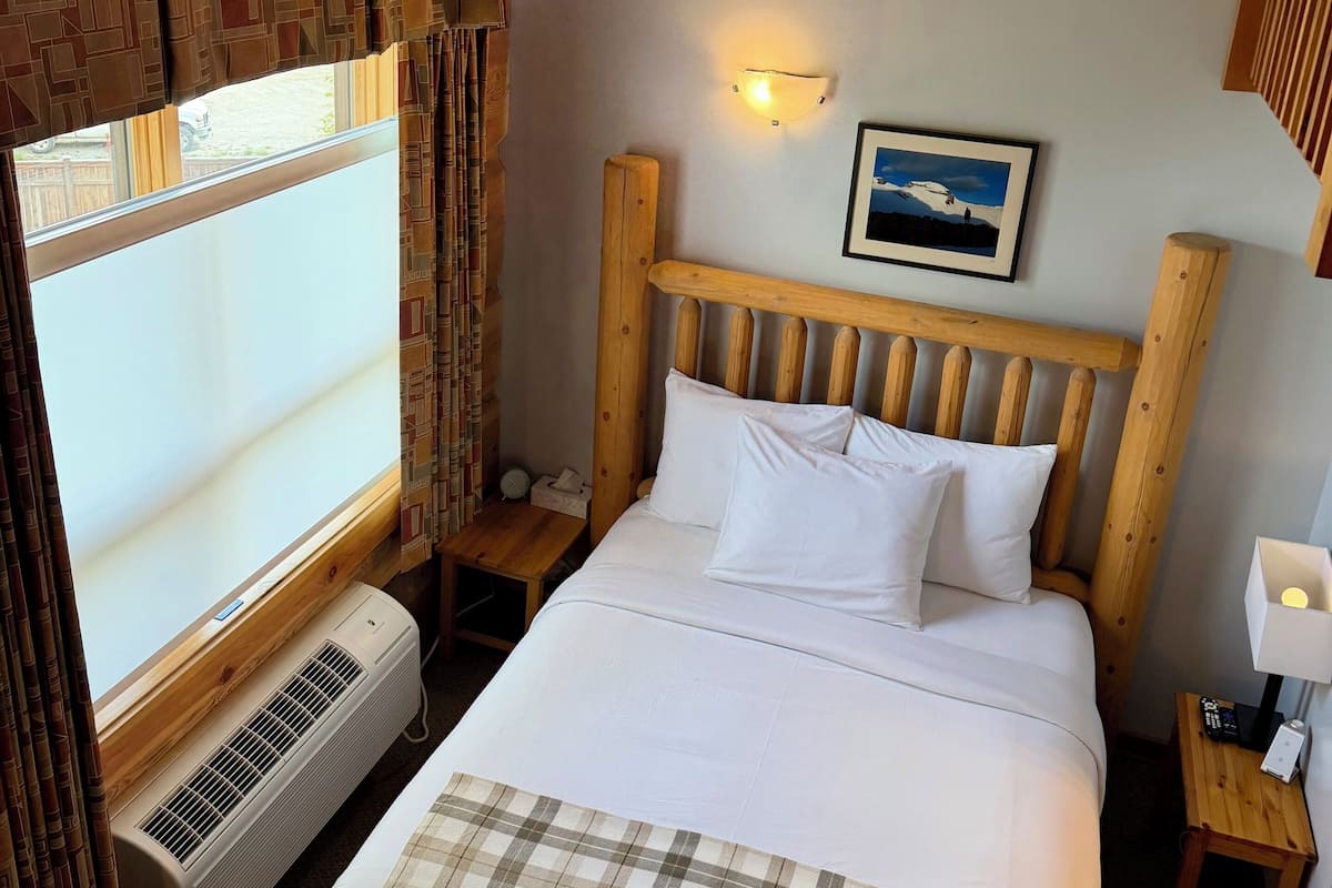 Habitación de hotel con cama, mesita de noche, luz de pared, cuadro enmarcado y ventana con cortinas.