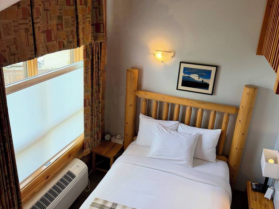 Habitación de hotel con cama, mesita de noche, luz de pared, cuadro enmarcado y ventana con cortinas.