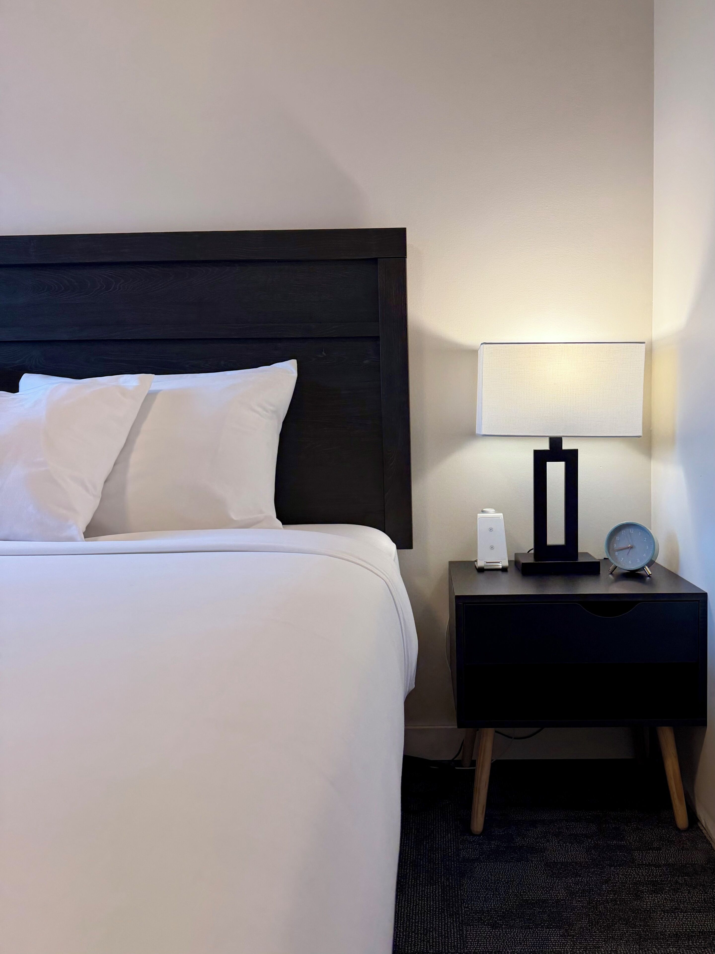 Un lit bien fait, avec du linge de lit blanc, une tête de lit sombre, une table de chevet avec une lampe, une horloge et un petit objet blanc.