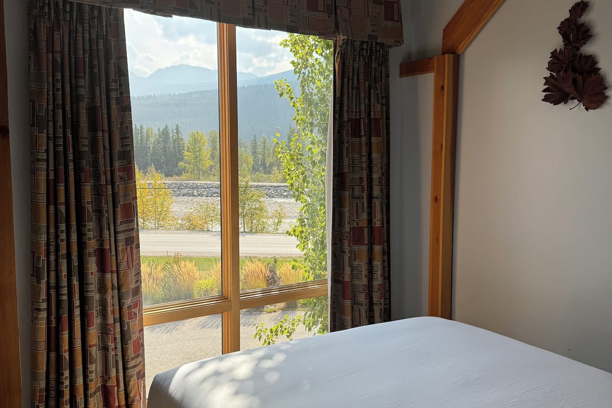 Un dormitorio con un ventanal grande, una cama y vistas a montañas y una carretera.