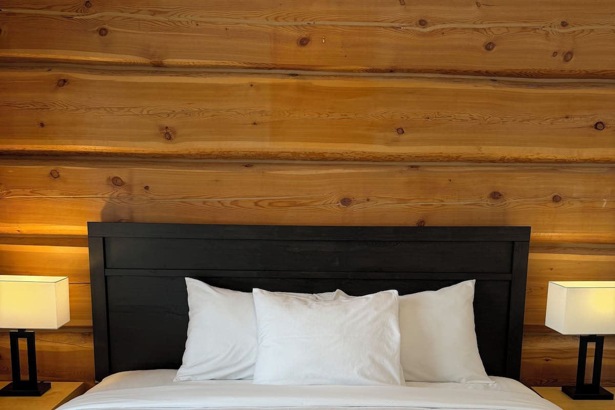 Una cama bien hecha con sábanas y almohadas blancas, un cabecero oscuro y dos lámparas de noche sobre mesitas de madera.