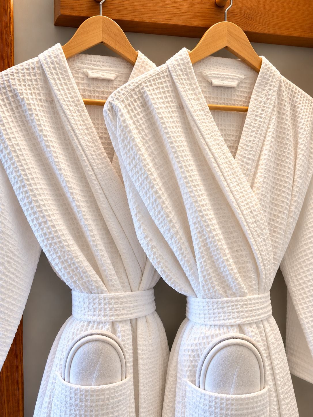 Two white bathrobes hanging on wooden hangers.