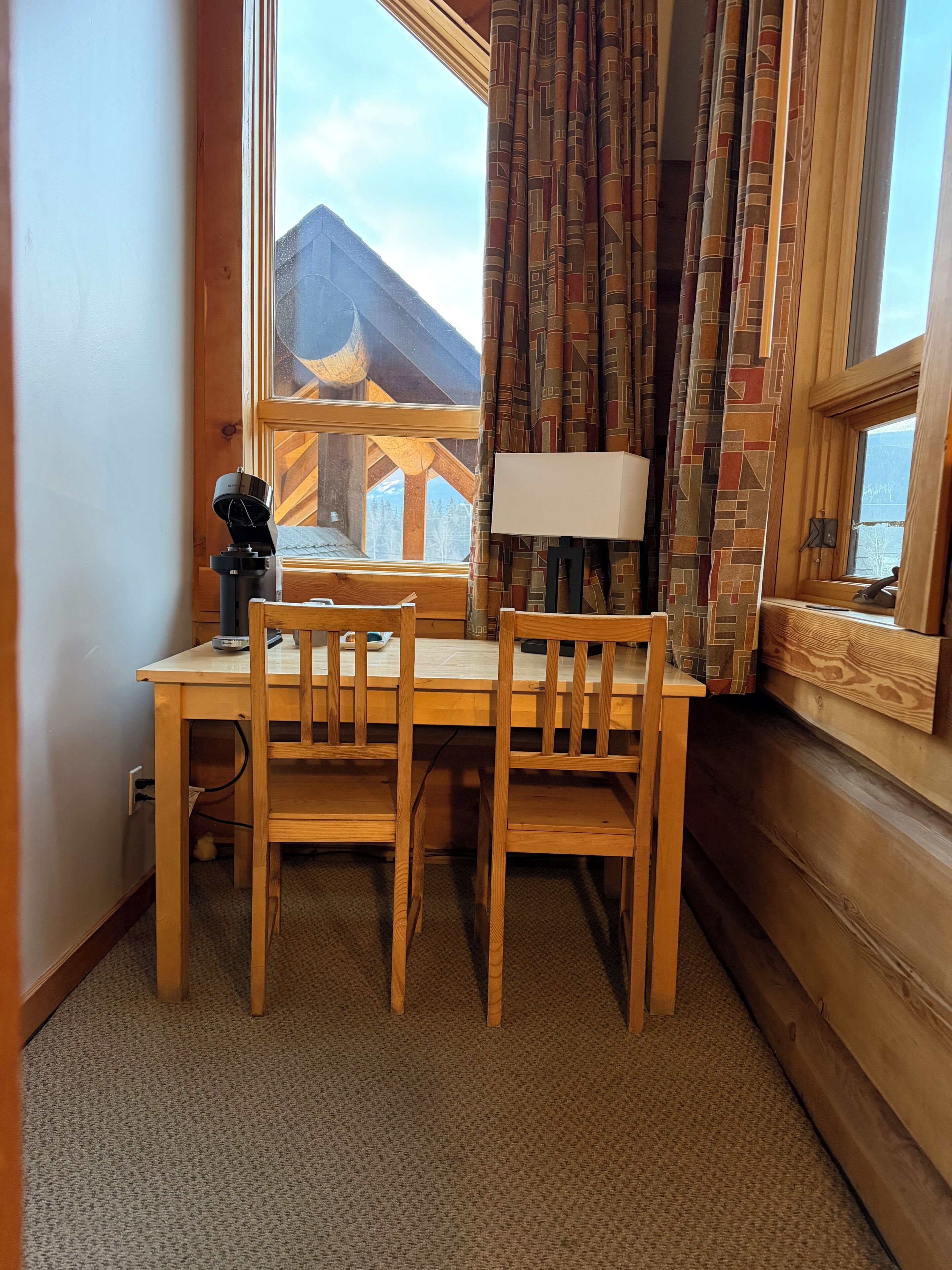 A wooden desk with two chairs, a lamp, and a camera in a room with a view of mountains through a window.