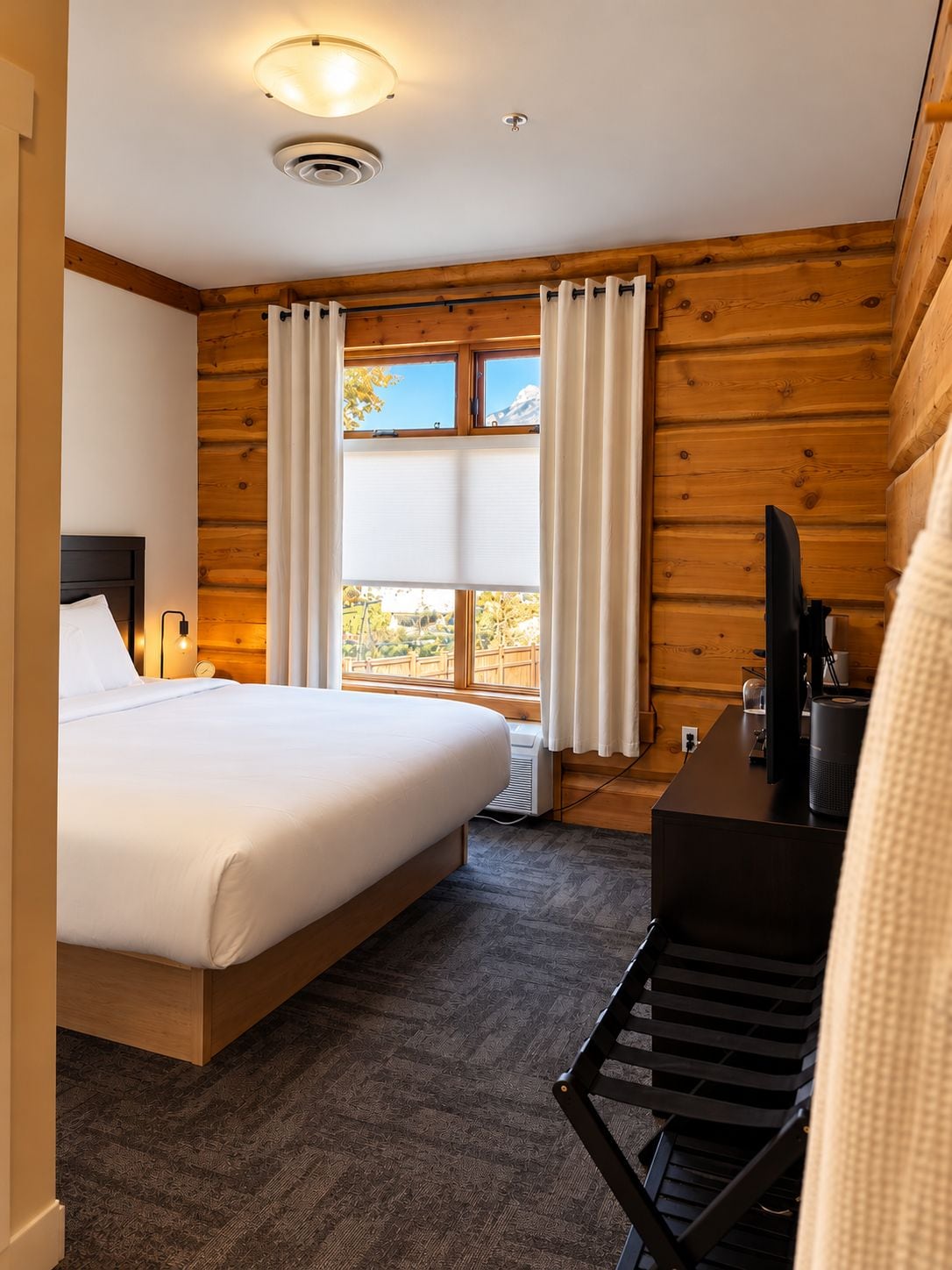 A hotel room with a bed, a desk with a computer, a window with curtains, and a wooden wall.
