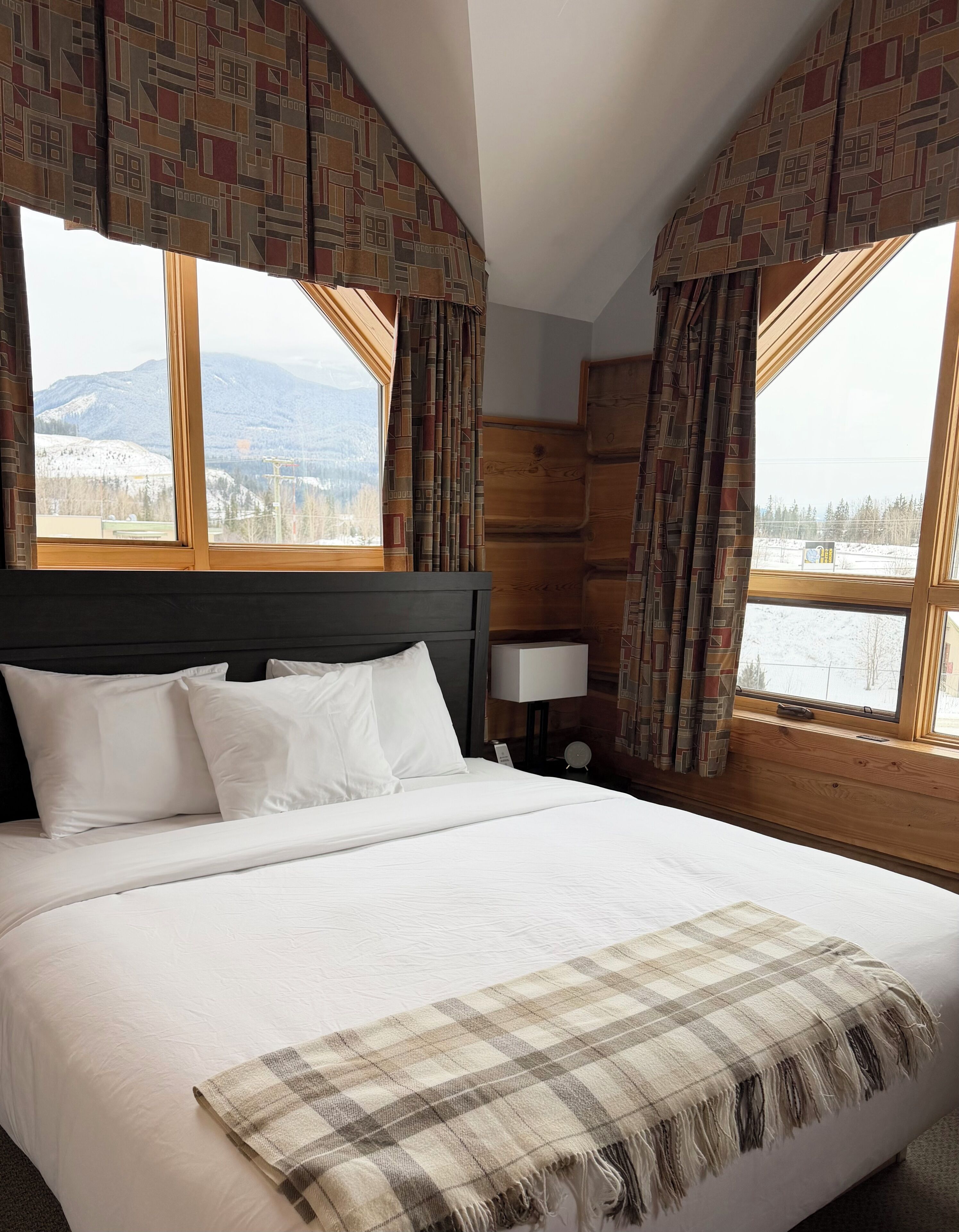 Habitación de hotel con cama, mesita de noche, lámpara y vistas a un paisaje nevado a través de amplios ventanales.