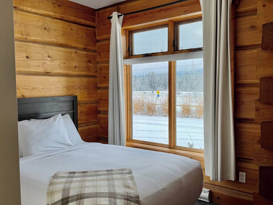 Een houten kamer met een bed, een raam met gordijnen en uitzicht op een besneeuwd landschap.