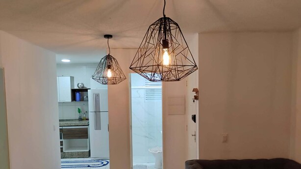Una sala moderna con un sofá oscuro, una mesa de centro de vidrio y dos lámparas colgantes geométricas. Se ve una cocina con armarios blancos y un refrigerador a través de una puerta abierta.