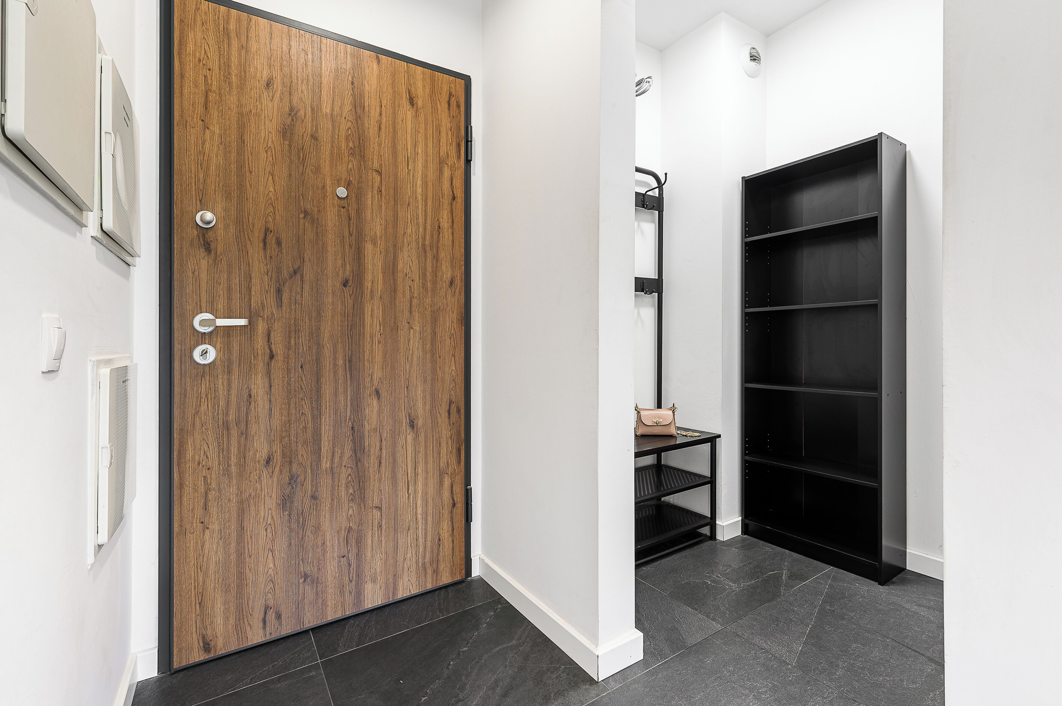 Un pasillo con una puerta de madera, una estantería negra y una pared blanca.