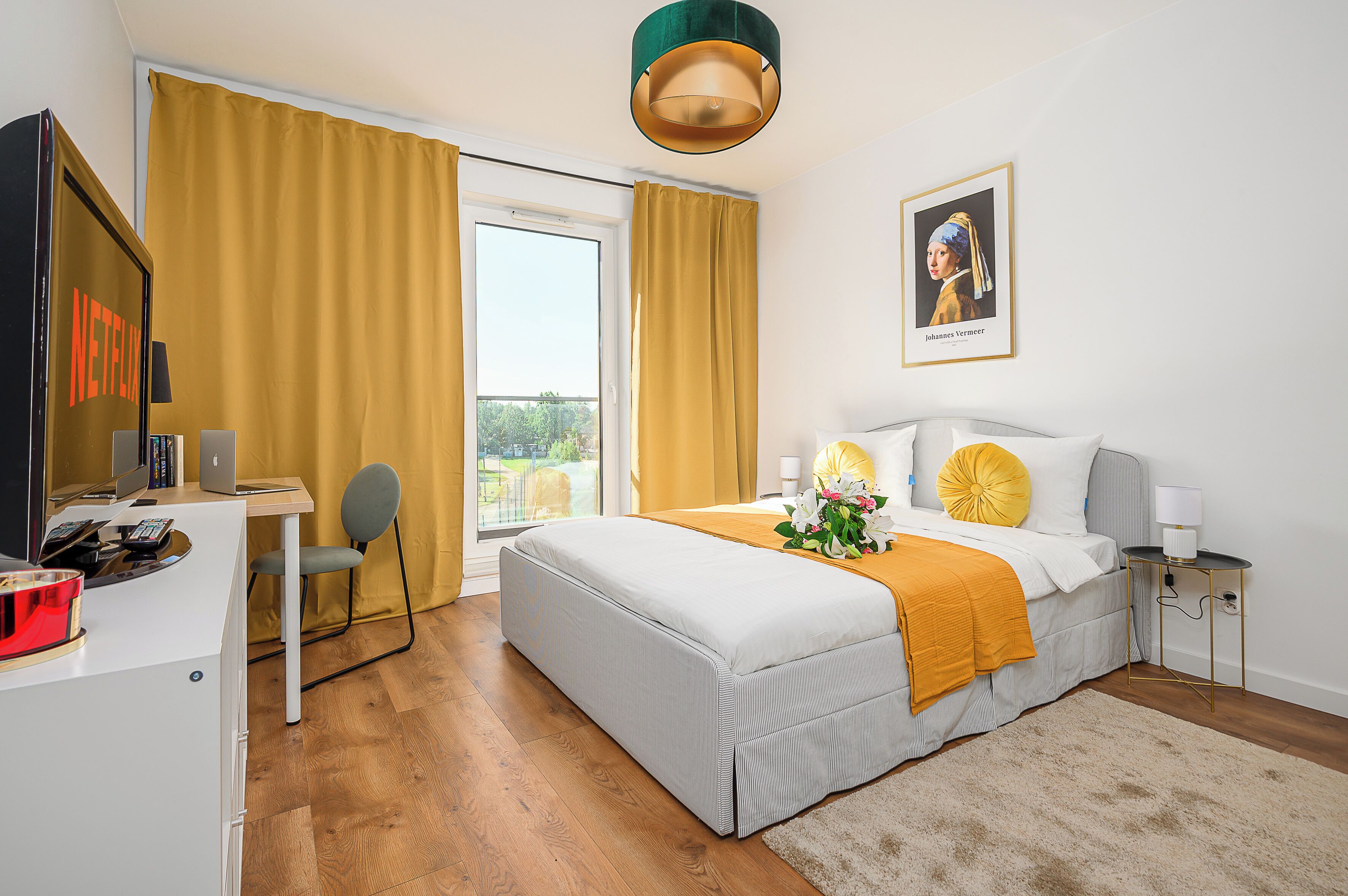 Une chambre d’hôtel avec un lit, un bureau avec un ordinateur, une chaise, une fenêtre avec des rideaux, un tableau au mur et un tapis sur le sol.