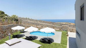 A pool area with lounge chairs and umbrellas, surrounded by a stone wall and a view of the sea. - Villa Calissea (Agia Pelagia)
