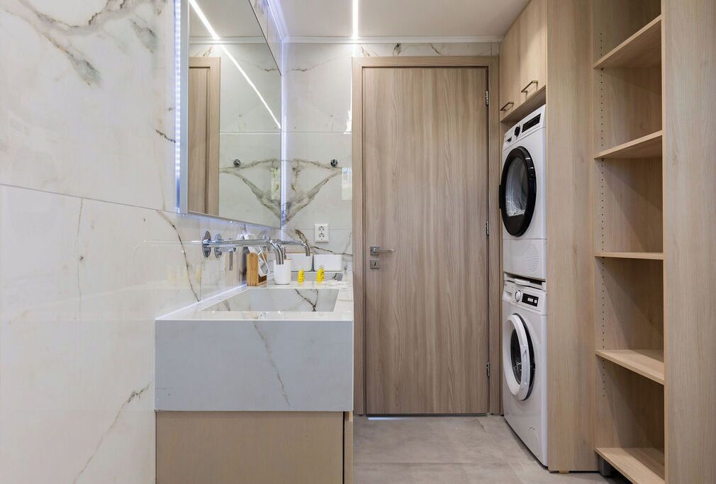 A compact bathroom with a marble vanity, a washing machine, and wooden shelving.