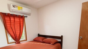 Un dormitorio con cama, ventana con cortinas, unidad de aire acondicionado y puerta de madera.