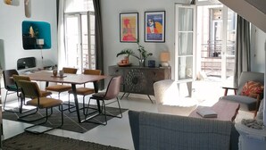 Un vaste espace de vie comprenant une table à manger, des chaises, un canapé et une table basse. Deux affiches encadrées ornent le mur, et une petite plante est posée sur une étagère.