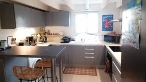 Une cuisine moderne avec des meubles gris, un réfrigérateur en acier inoxydable, un four à micro-ondes et une machine à café. On y trouve deux tabourets de bar et un petit tapis sur le sol.