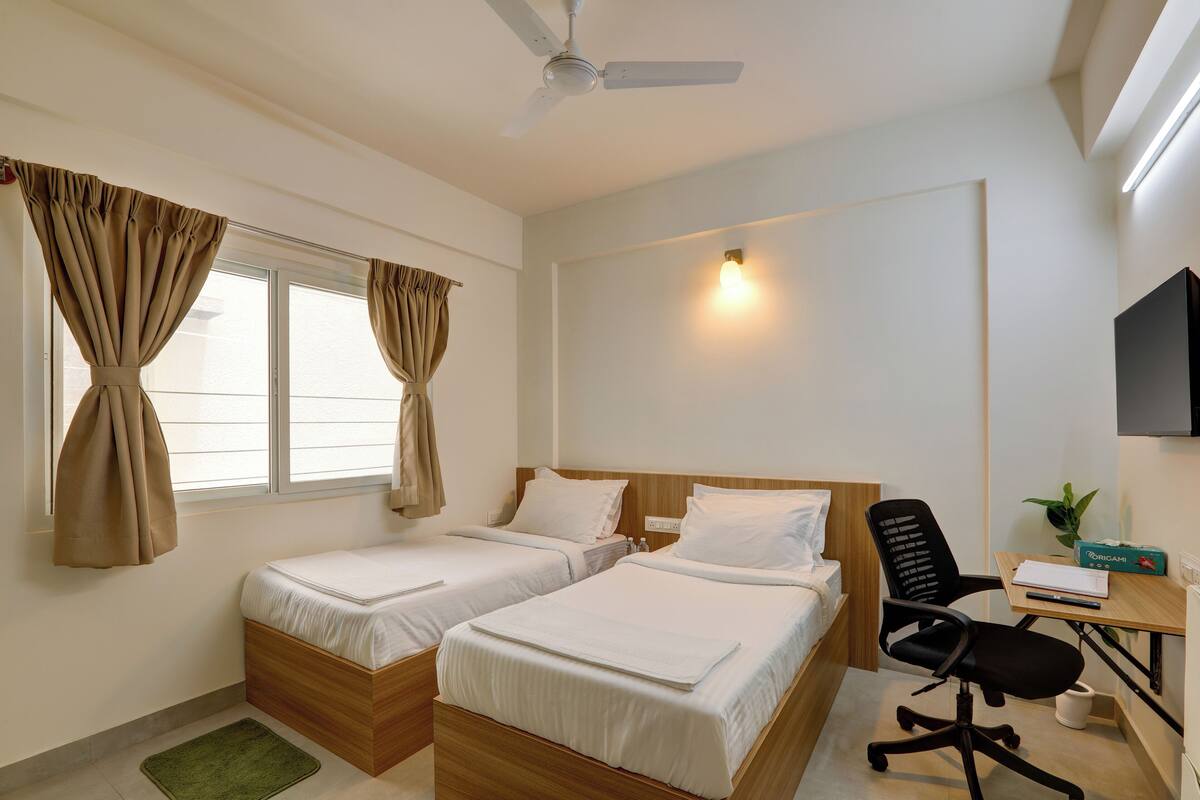 Habitación de hotel con dos camas individuales, un escritorio con silla, ventilador de techo y ventana con cortinas.