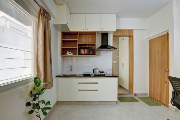 A compact kitchen with white cabinets, a microwave, and a sink. There's a wooden shelf above the counter and a green plant on the left.