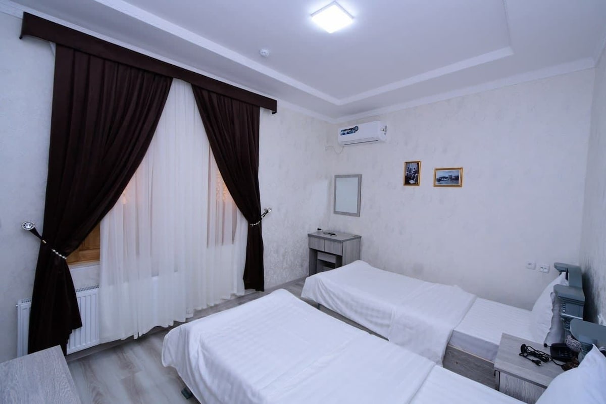 A hotel room with two beds, a small bedside table, a mirror, and a radiator.