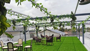 Ein Außenbereich mit Sitzgelegenheiten, ausgestattet mit Kunstgrras, Holzkreislern und -tischen, unter einer Pergola mit hängenden Pflanzen und Laternen.